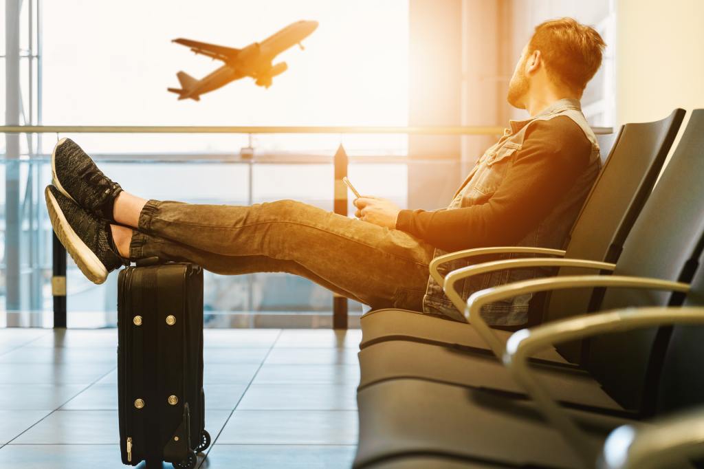 A young man in tennis shoes waiting in an airport terminal at sunset with his feet propped on his suitcase as a plane takes off outside. 
